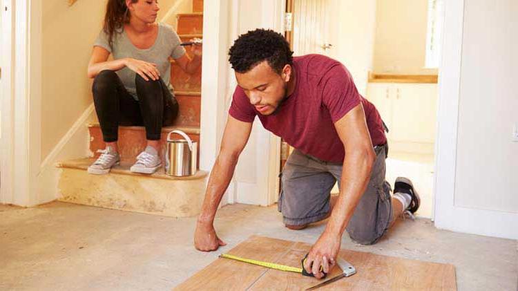 一名男子正在测量地板上的一块木板，而一名女子正在粉刷墙壁，以帮助完成他们的DIY家居装修.